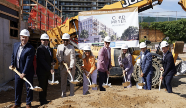 Eight people in hard hats hold shovels during a groundbreaking ceremony in front of construction machinery and a banner for Cord Meyer Development.