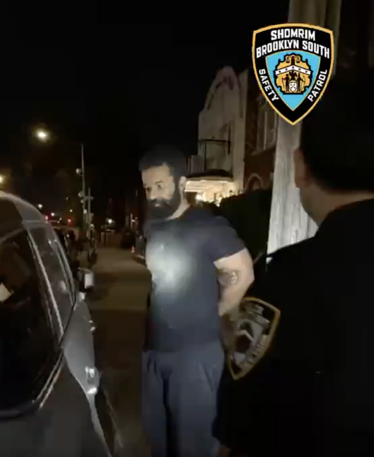 Man with a beard being arrested by an officer at night.