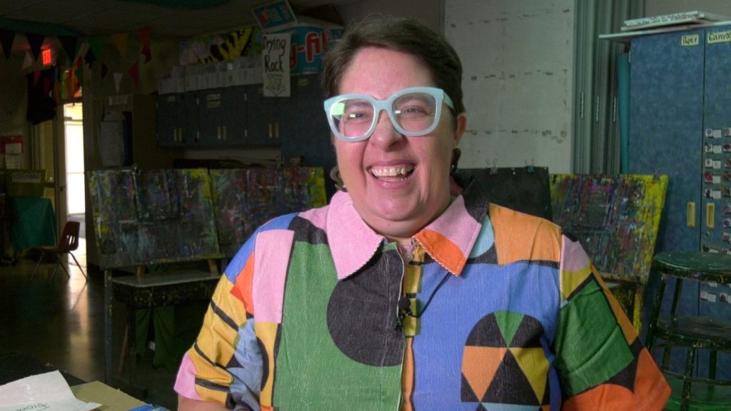 A woman with blue glasses and a rainbow collared dress laughs