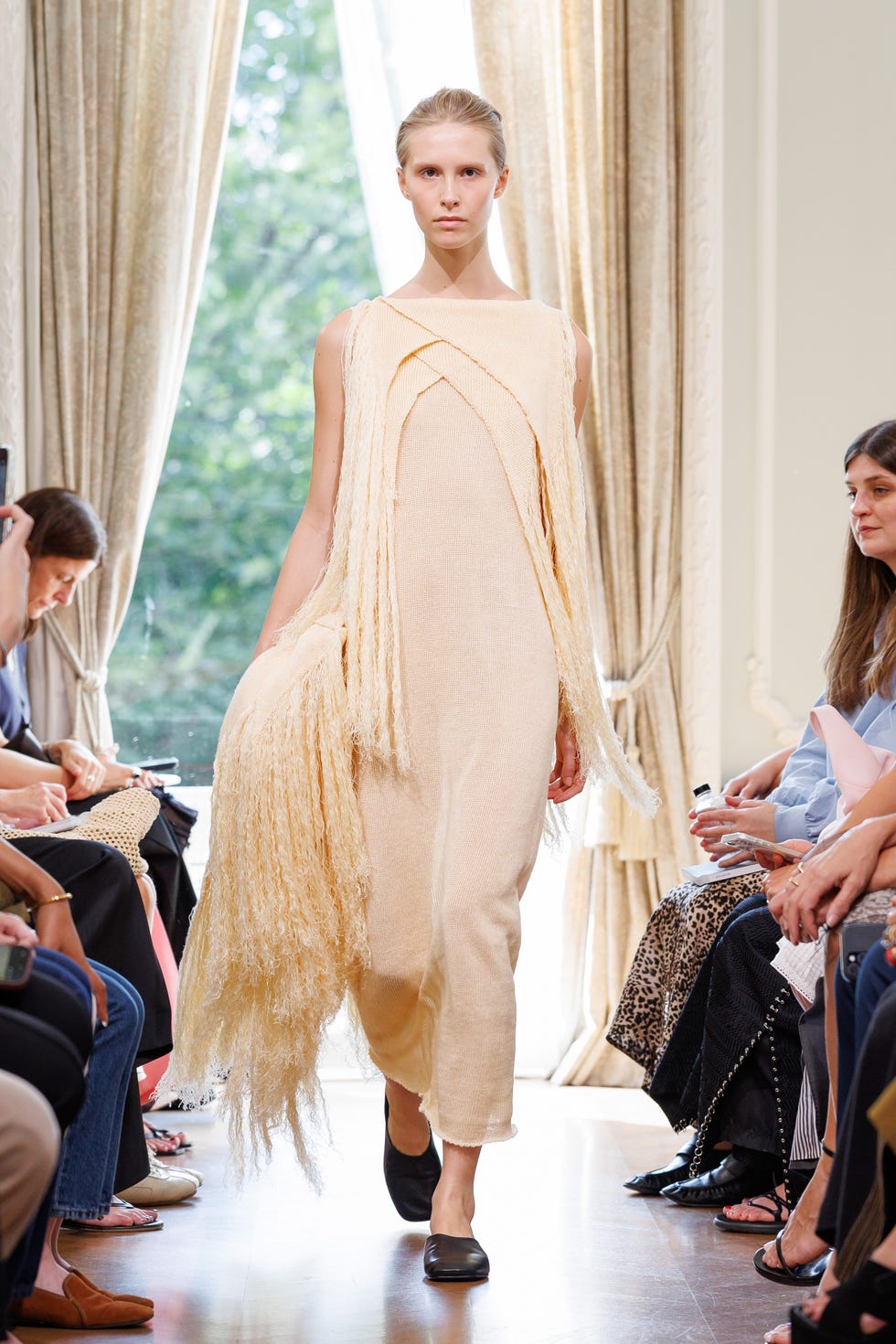A model walks the runway in a beige dress with fringe detailing during a fashion show.