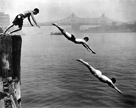 Three young men diving into a river from a wooden dock, with a bridge visible in the background.
