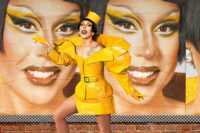 A drag queen in a yellow latex outfit, with one arm in the air.