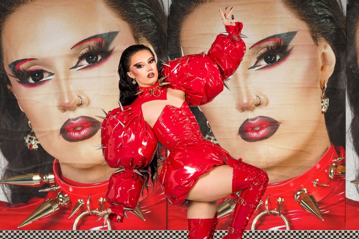 A drag queen in a red latex outfit slightly lent to one side with one arm in the air.