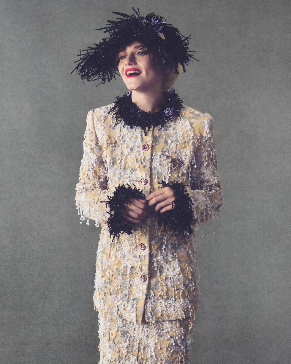 Julia Garner wearing a textured suit with purple accents and a large decorative hat, smiling against a gray background.