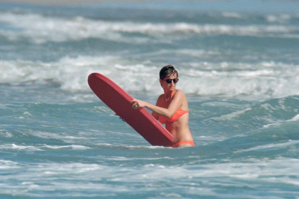 Princess Diana in orange two-piece swimsuit and sunglasses standing in the sea holding a red bodyboard 