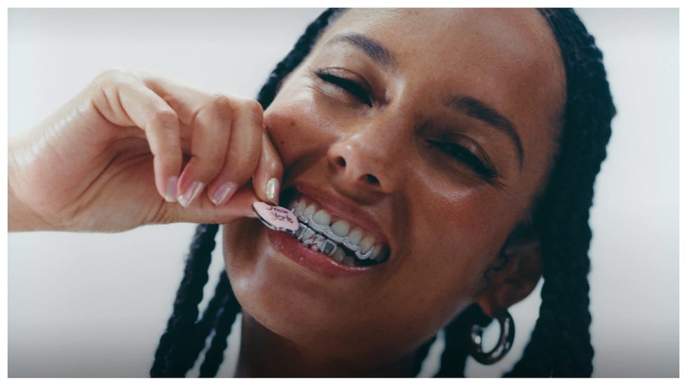 A person with braided hair smiling while brushing their teeth with a pink toothbrush.