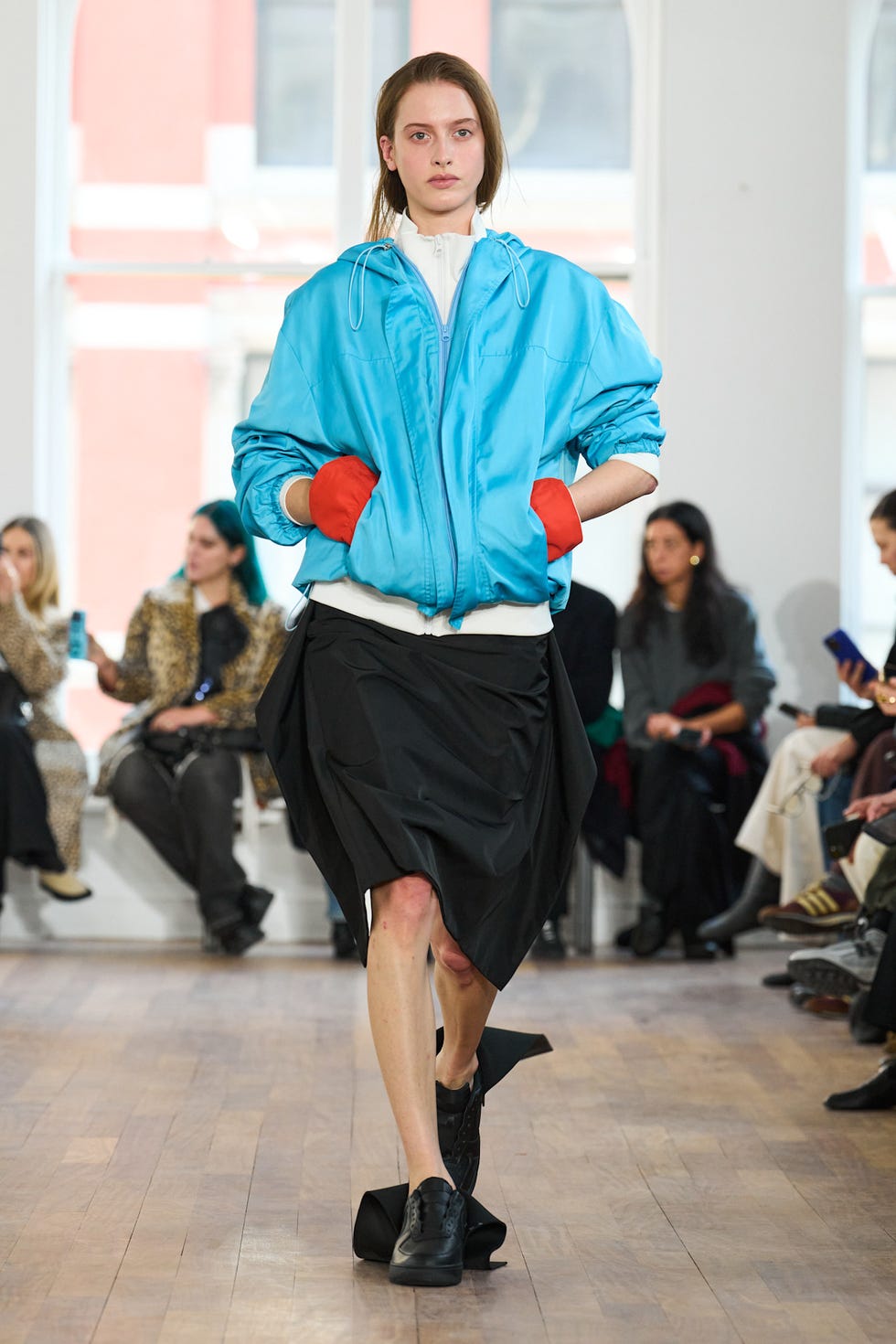 Model walking in a fashion show wearing a blue jacket, black skirt, and red gloves, with an audience in the background.