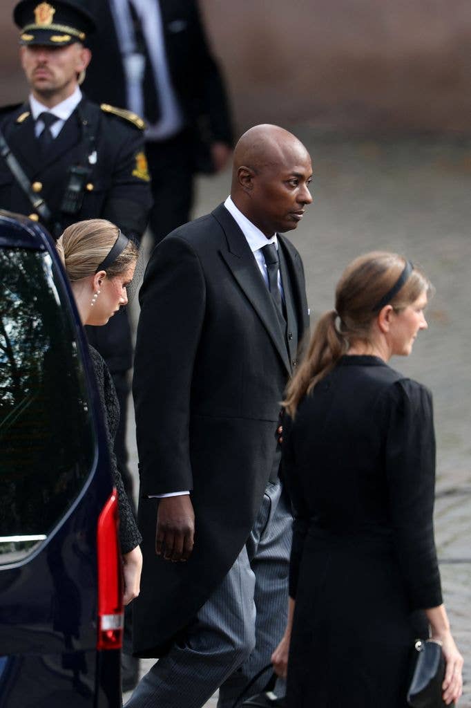 Man in black suit and tie walks with two women in black, one holding a purse.