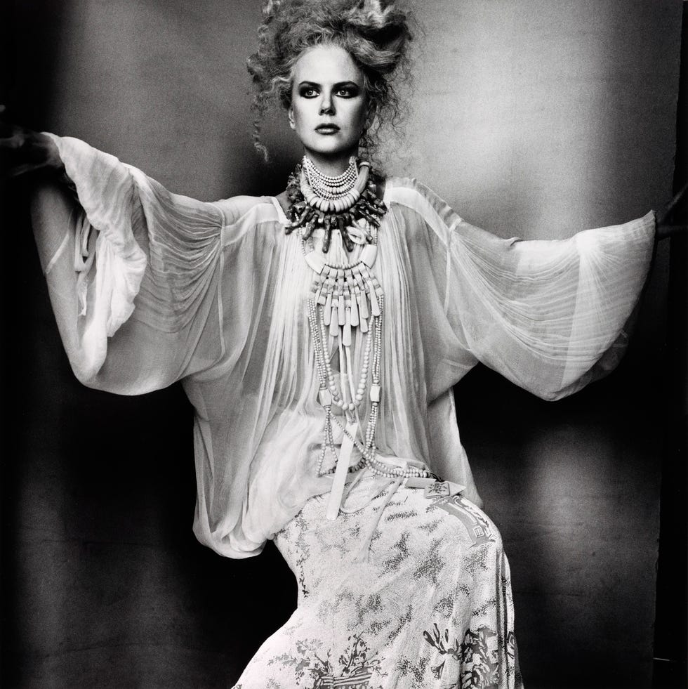 A model wearing bold jewelry and a flowing white garment, striking a dramatic pose in black and white photography.