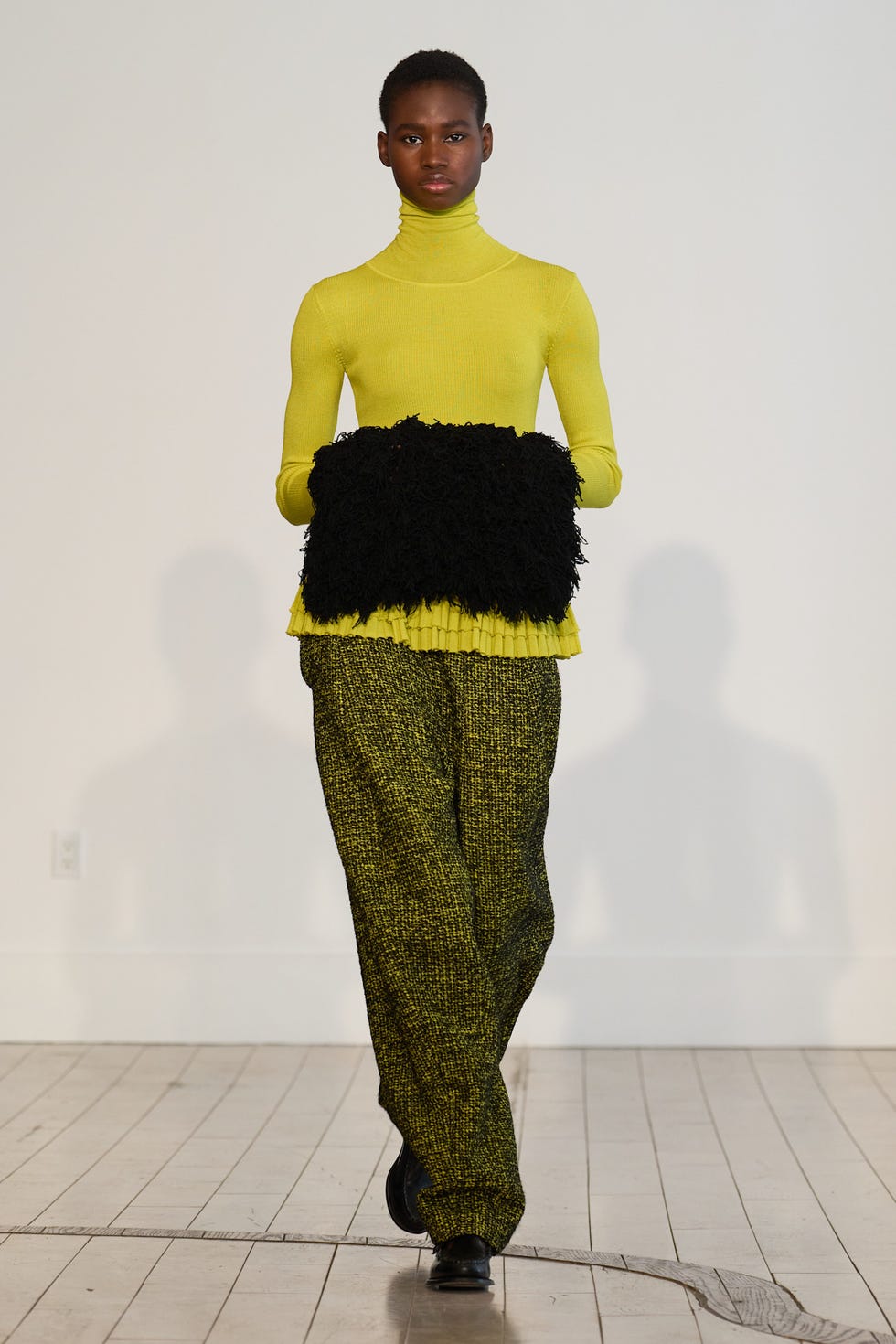 Model walking on runway in vibrant yellow turtleneck, black textured clutch, and patterned green pants.