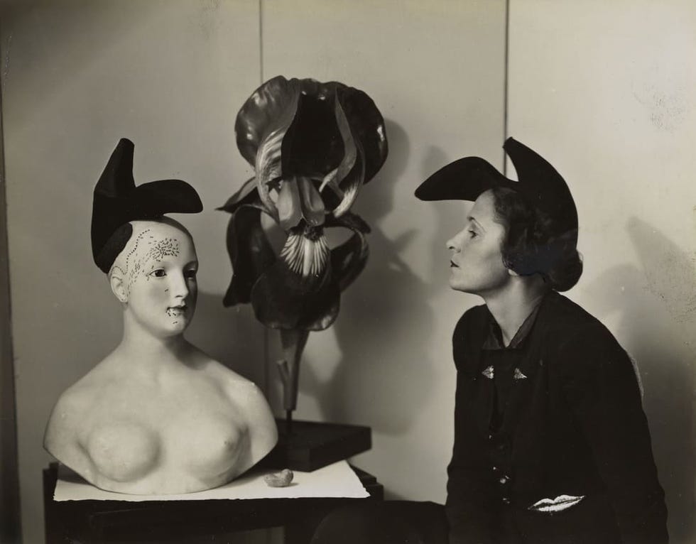 A mannequin wearing a hat sits beside a woman in profile, both positioned near a decorative plant.