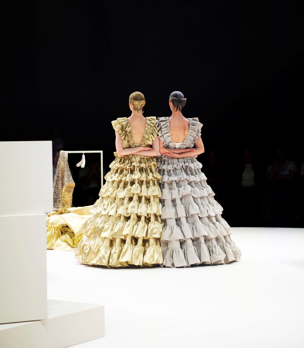 Two models in dramatic, ruffled gowns, one gold and one gray, pose back-to-back on a fashion runway.