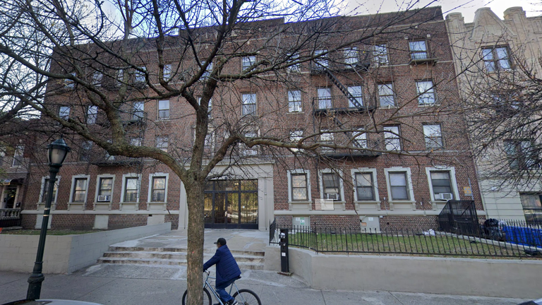 A person rides a bicycle on the sidewalk in front of a large brown brick apartment building with multiple windows and a tree in the foreground.