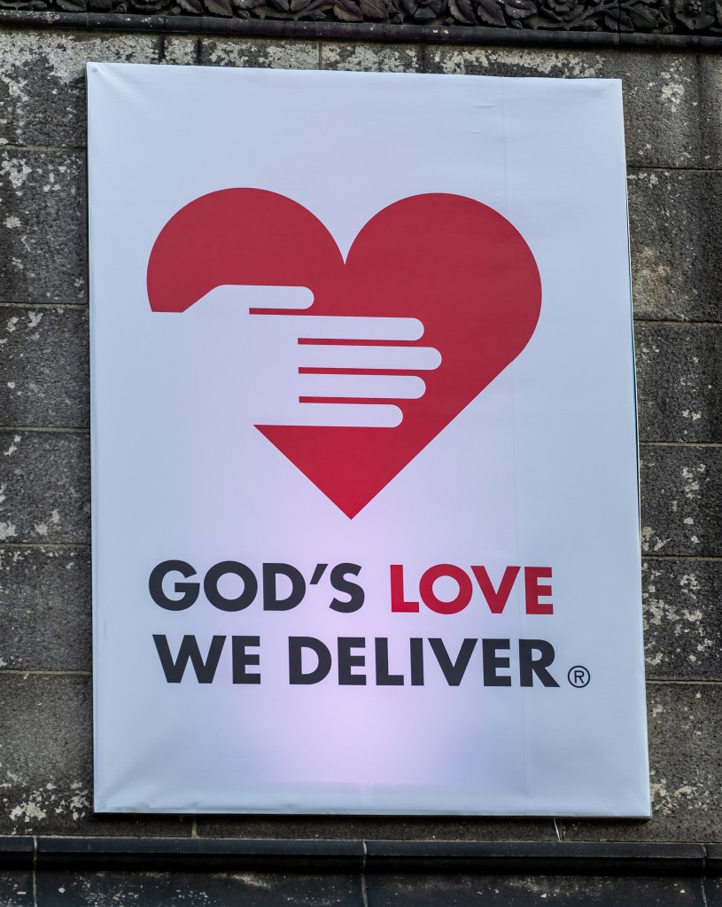 A banner for God's Love We Deliver featuring its logo, a red heart embraced by a white hand, mounted on a stone wall at the Cathedral of St. John the Divine.