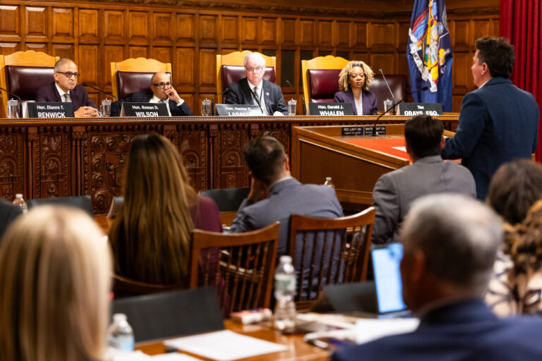 A formal hearing takes place in a wooden-paneled chamber, with four officials seated at a long table and a person standing to speak before them.
