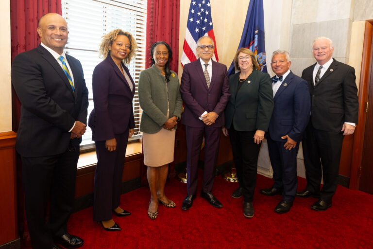 Seven formally dressed individuals pose together in an official room with American and state flags, standing on a red carpet.
