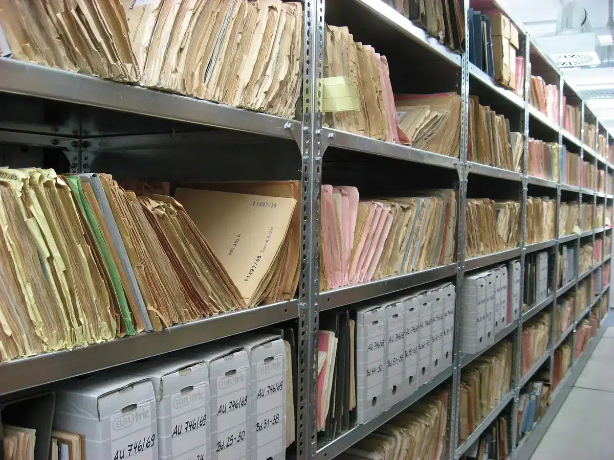 empty municipal records room with rows of unmarked boxes and stacks of paper, a single laptop open on a table showing an open document portal