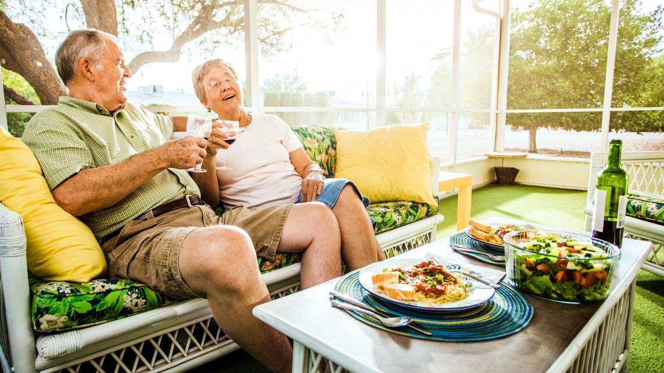 An elderly man and woman sit on a white wicker couch in a bright sunroom or screened porch, holding glasses of red wine and laughing joyfully. A white table in front of them holds plates of pasta with sauce, a clear bowl of salad, and a green wine bottle. The couch features green floral cushions and yellow pillows, while bright sunlight streams through the screened windows, revealing green trees in the background.