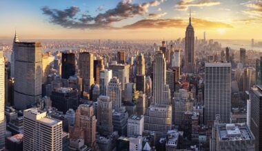 An aerial panorama of the New York City skyline at sunset. Numerous tall skyscrapers, including the Empire State Building, dominate the landscape. The sky is a gradient of blue, orange, and yellow, with scattered clouds illuminated by the setting sun. The city stretches into the distance, with water visible towards the horizon.