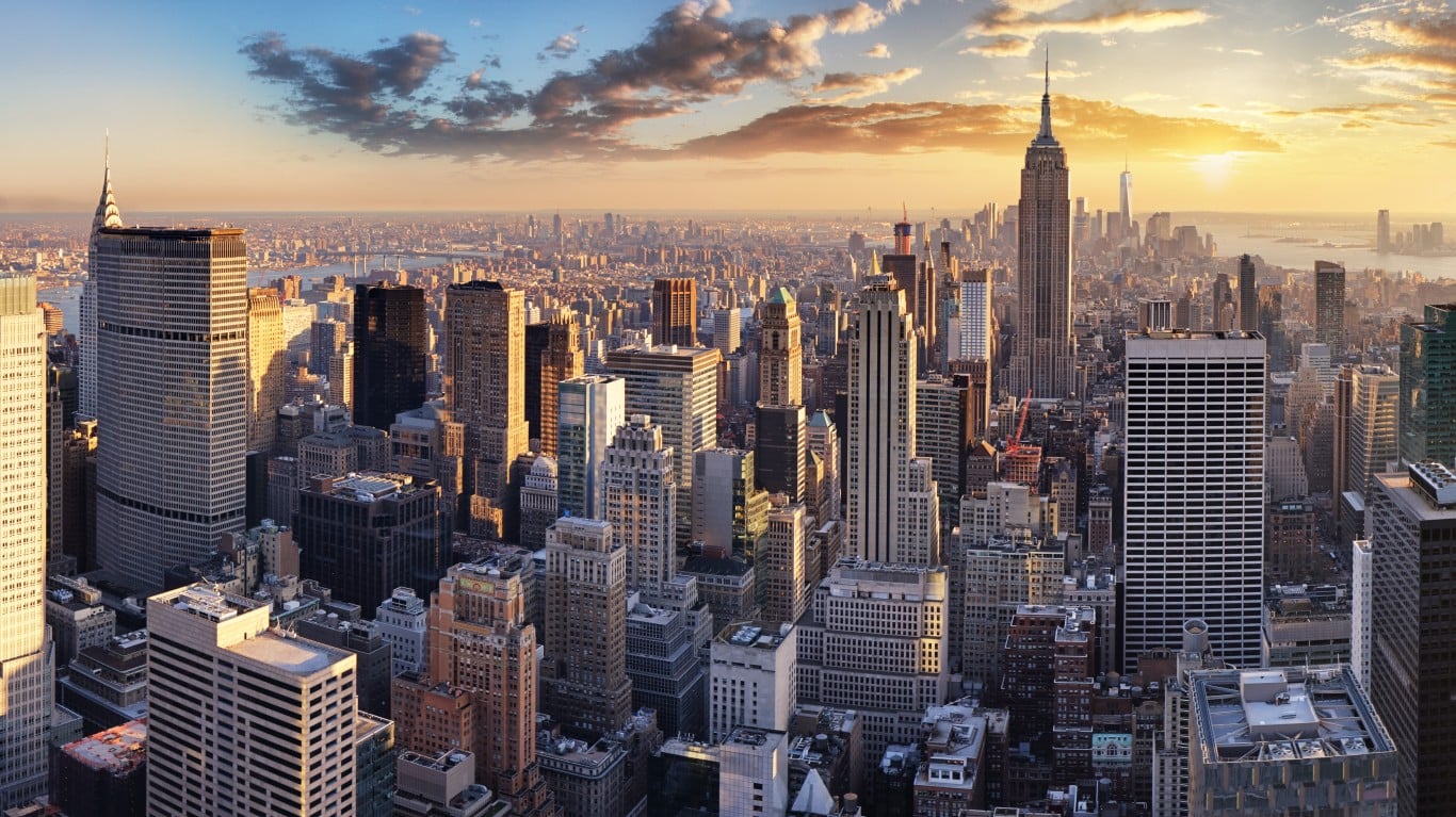 An aerial panorama of the New York City skyline at sunset. Numerous tall skyscrapers, including the Empire State Building, dominate the landscape. The sky is a gradient of blue, orange, and yellow, with scattered clouds illuminated by the setting sun. The city stretches into the distance, with water visible towards the horizon.