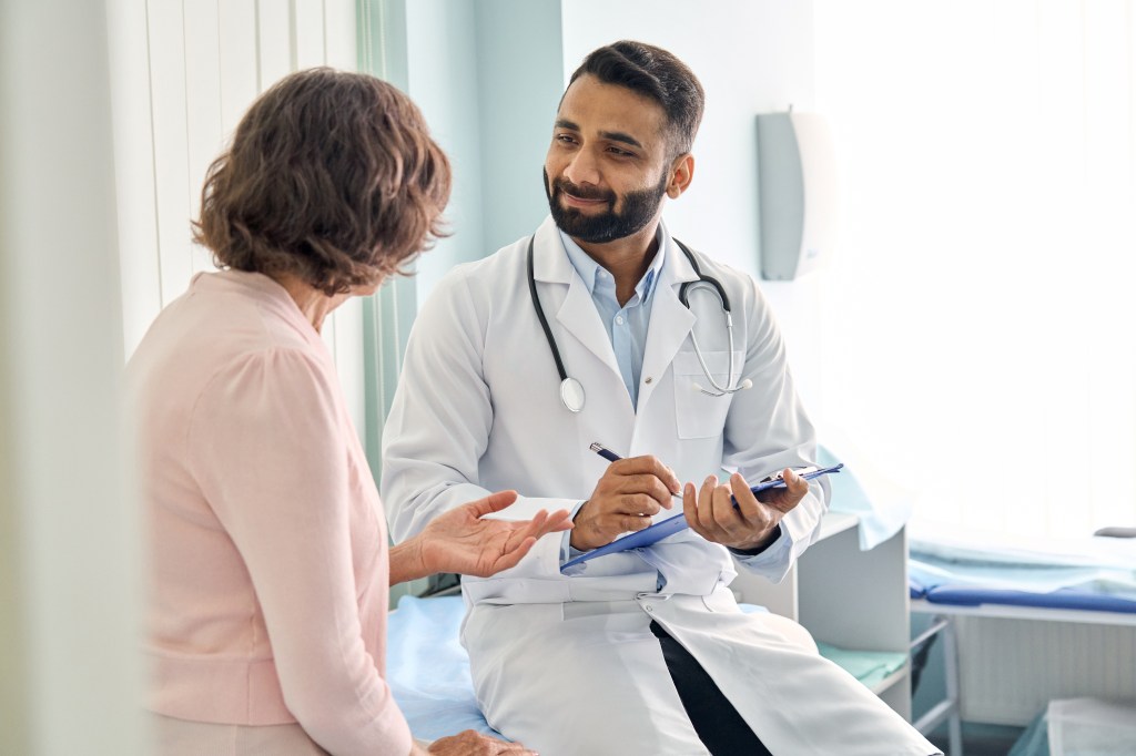 An Indian male doctor consulting a senior patient while filling a medical form.