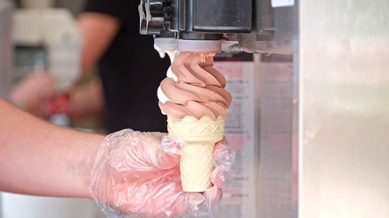Soft-serve chocolate ice cream is dispensed from a machine into a cone.