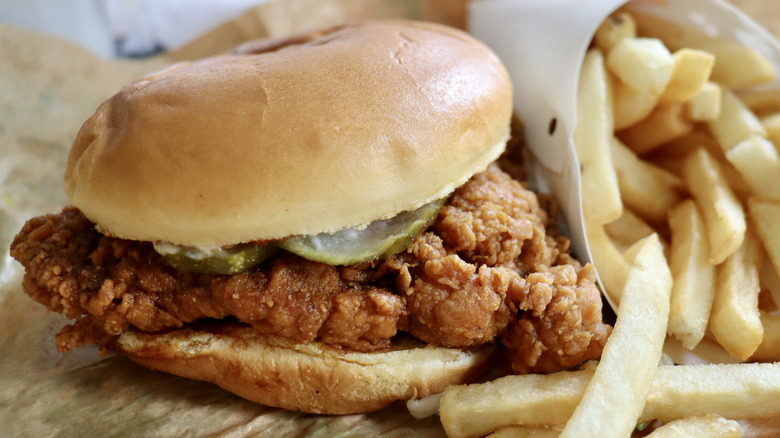 A fried chicken sandwich with pickles sitting on paper, beside a cardboard sleeve of fries