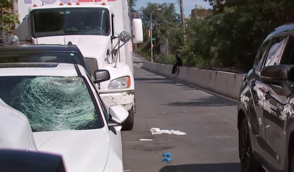 A white car with a shattered windshield, a white truck, and a dark car parked on a road, with what appears to be discarded items on the pavement, and a person sitting on a wall in the background.