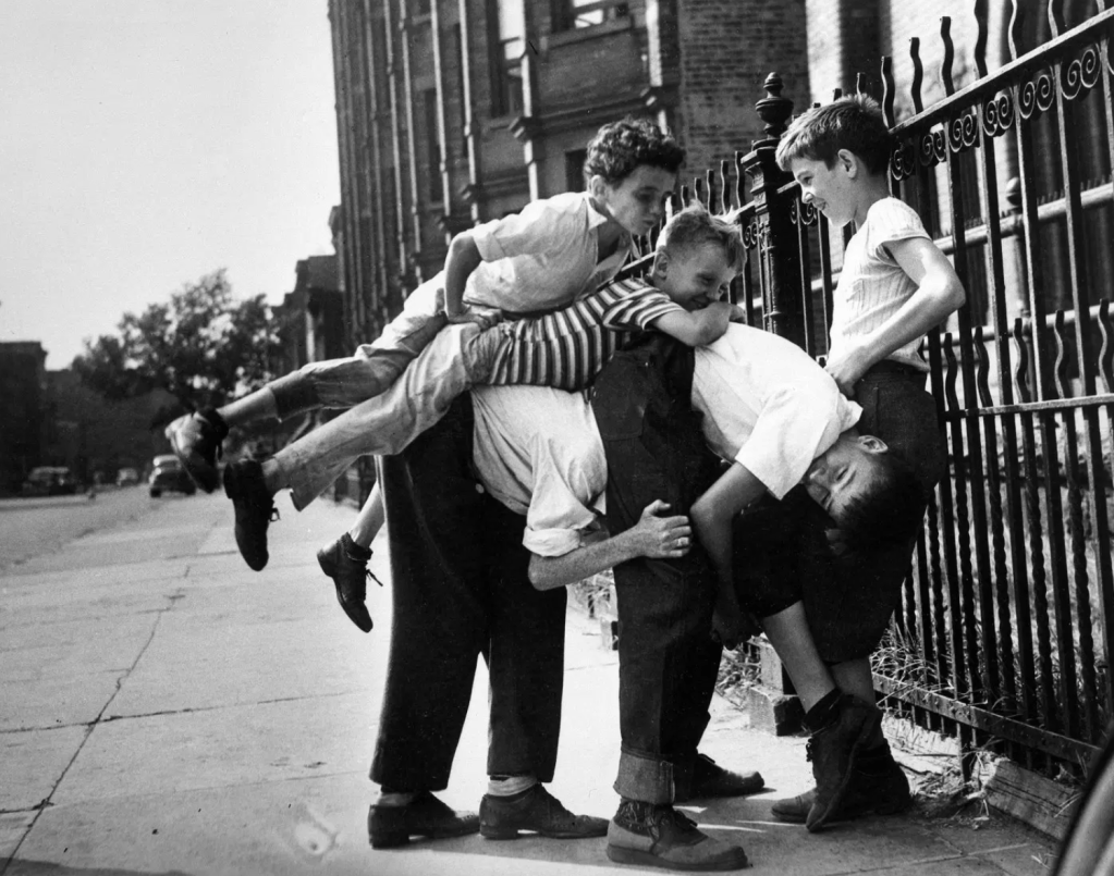 A group of children playfully climbing on each other near a street and a wrought-iron fence, capturing a moment of childhood fun in black and white.