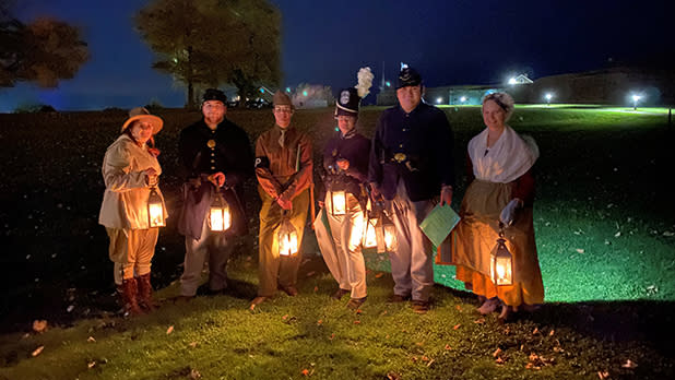 Six people in historical costumes pose at night in a grassy field, holding glowing lanterns; distant lights and trees create a warm, festive mood during a Lantern Tour at Fort Ontario.