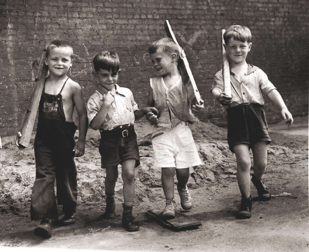 Four young boys walking together, smiling and holding wooden sticks. They are dressed casually, with varying styles of shorts and shirts, in a dusty outdoor setting by a brick wall.