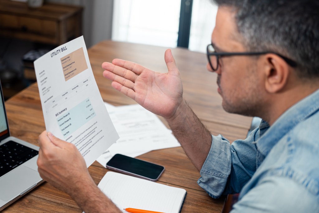 A man in glasses holds up a utility bill with a large sum and gestures in frustration.