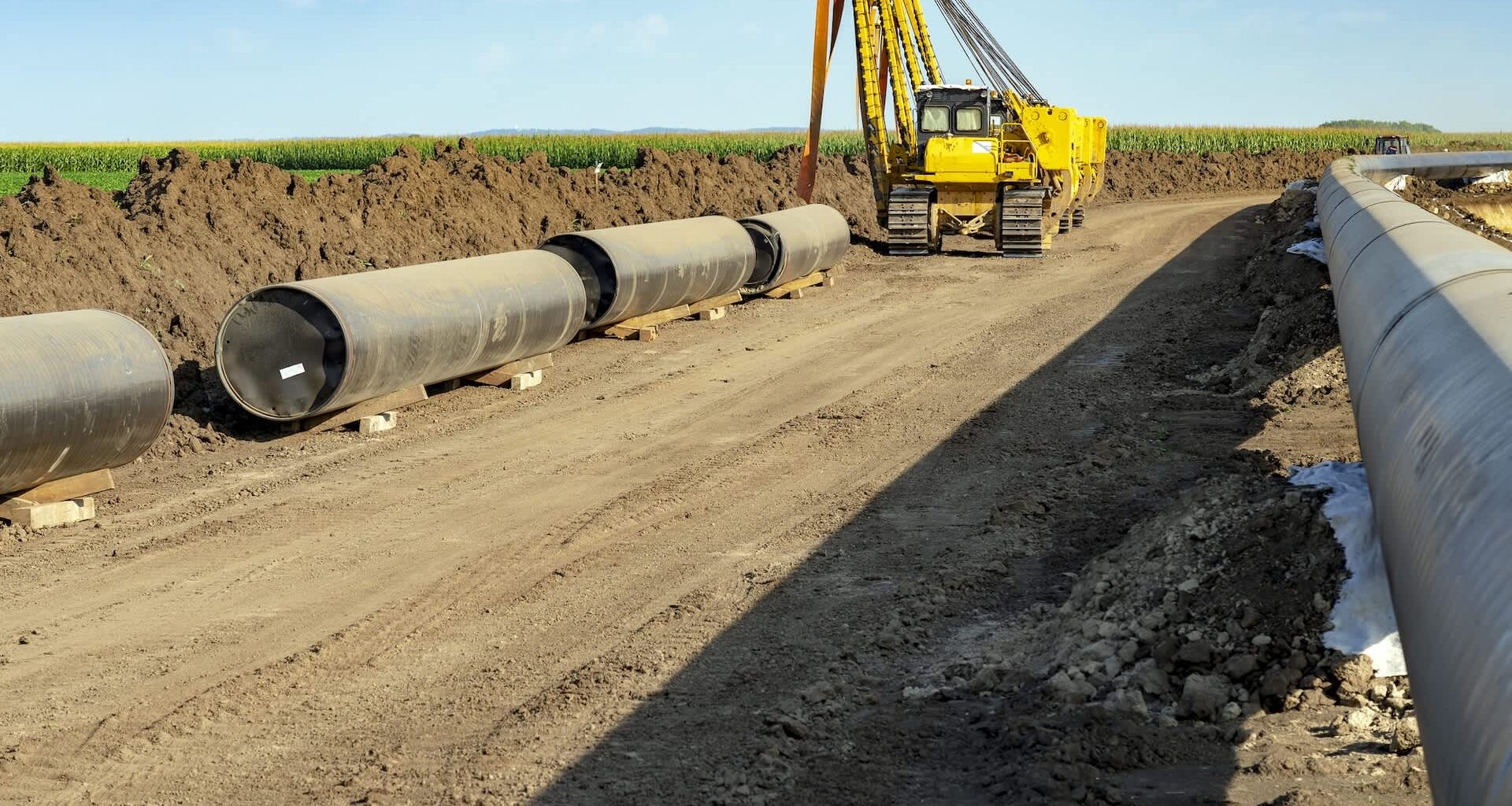 Heavy machinery is working on a construction site with large pipes lined along a dirt path near agricultural fields.