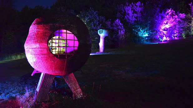 Nighttime outdoor light installation: a glowing spherical sculpture on legs, with colorful illuminated trees and a smaller orb in the background during Night Lights at Griffis Sculpture Park.