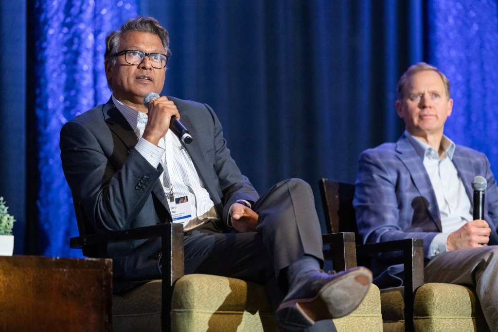 Dru Rai speaking into a microphone while sitting next to another man.