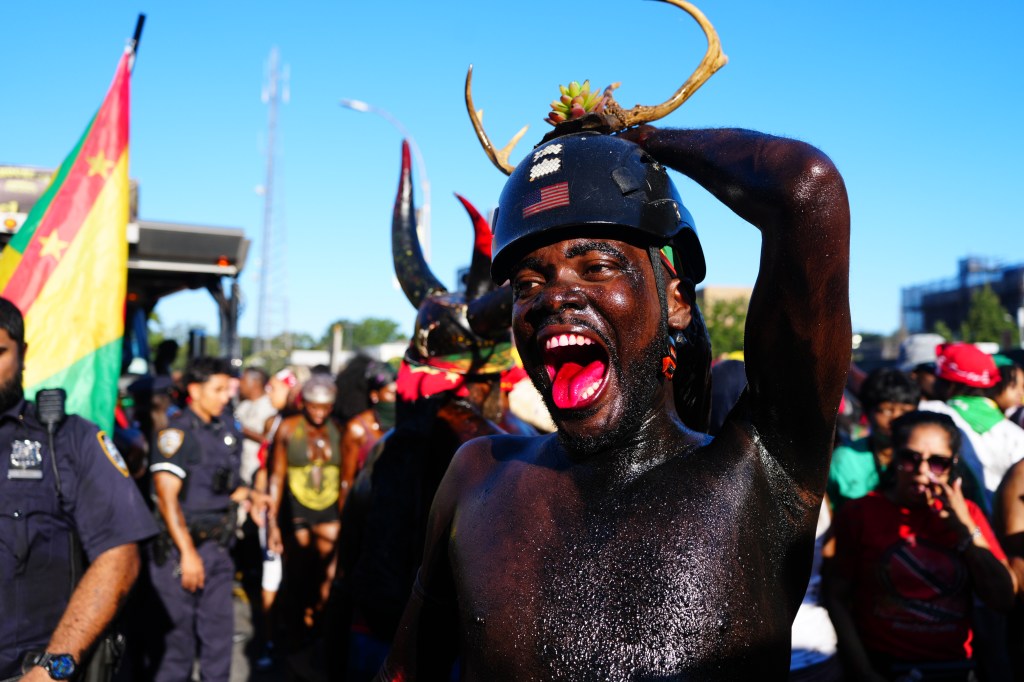 A person covered in black paint and wearing a helmet with antlers and a succulent on top, shouts during J'Ouvert celebrations in Brooklyn.