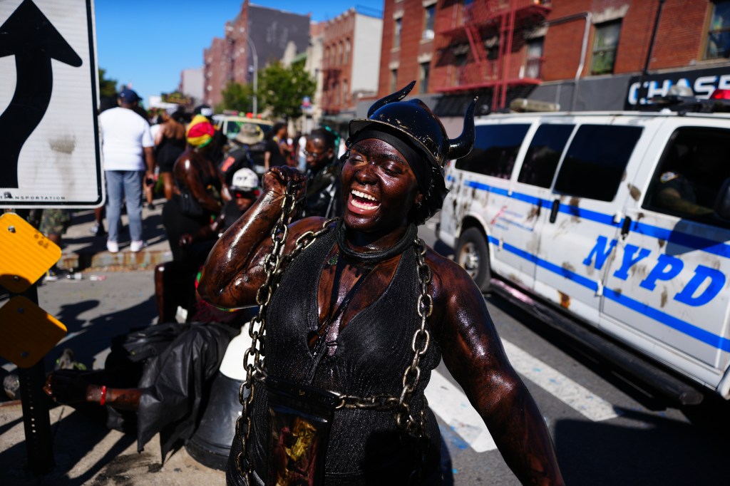 A participant in the J'Ouvert celebration, covered in dark paint and wearing a horned helmet, laughs and sings.