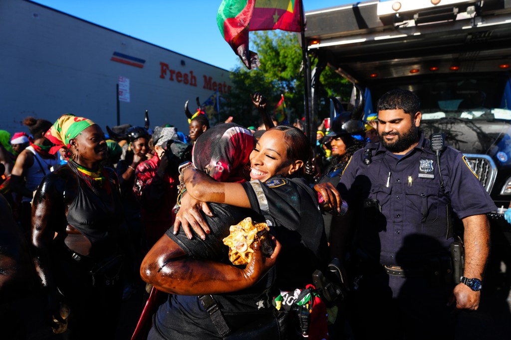 Two women covered in dark pigment embrace during J'Ouvert celebrations, with a police officer and other revelers in the background.