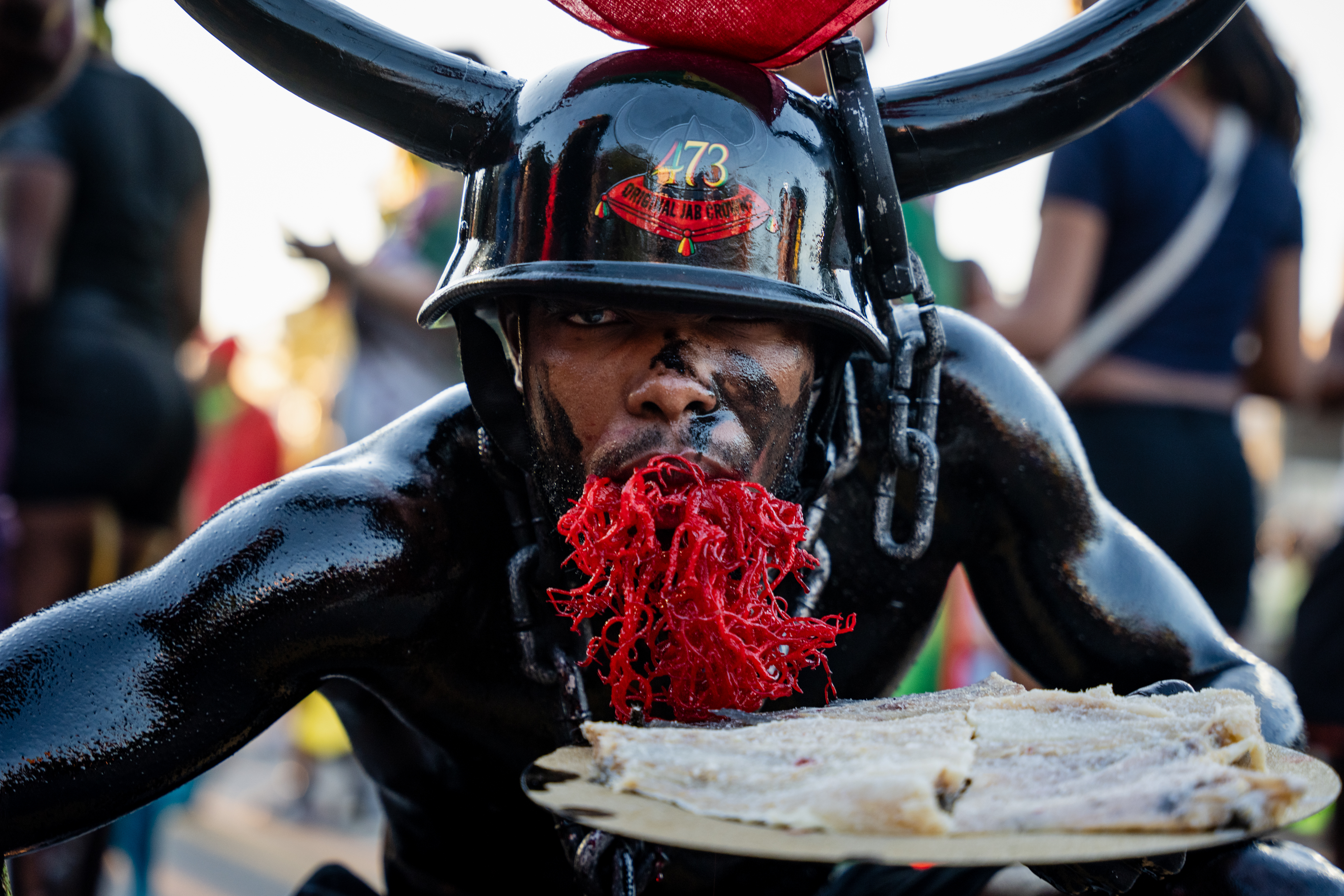 A person in black body paint and a horned helmet with red decoration around their mouth, holding a plate of dried fish, during J'Ouvert festivities.