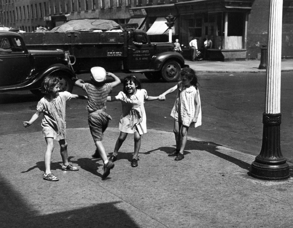 Four children playing together on a city street, two girls holding hands while a boy jumps in the center, with vintage cars and buildings in the background.