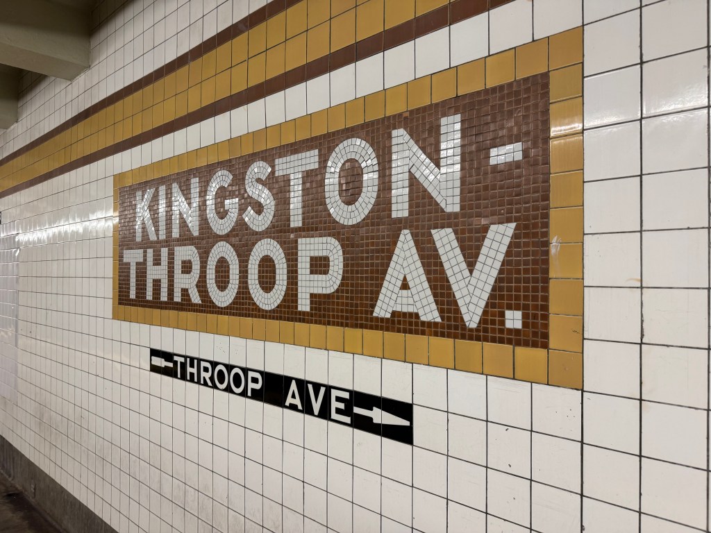 A brown, yellow, and white tiled subway sign for "Kingston-Throop Av." with an arrow pointing to "Throop Ave."