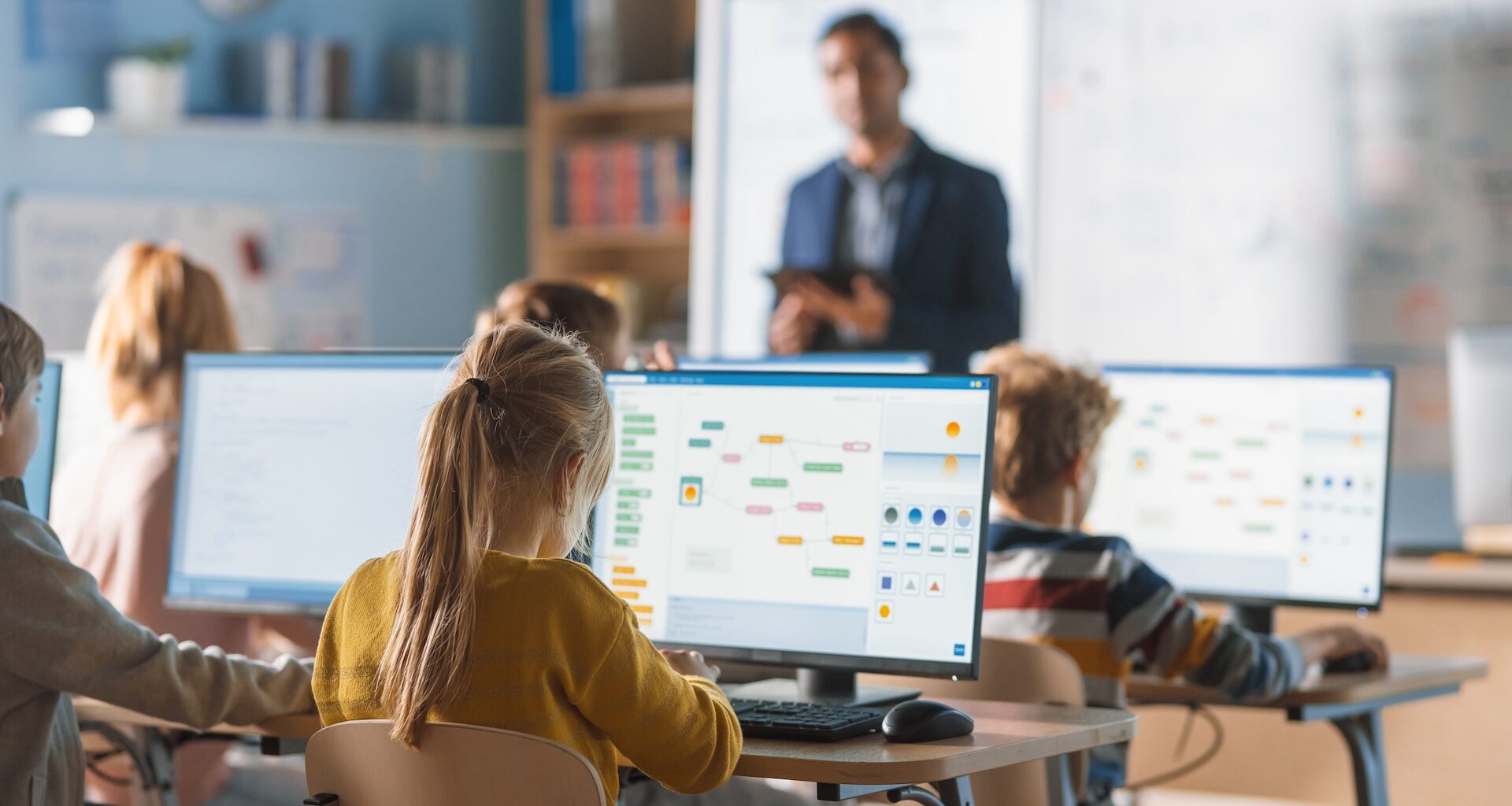 Students work on computers in a classroom while a teacher instructs in the background.