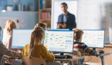 Students work on computers in a classroom while a teacher instructs in the background.