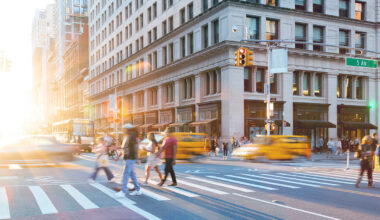 People cross a busy city street with blurred yellow taxis and sunlight streaming in. A large building and traffic lights are visible near the intersection of 5th Ave, capturing the vibrant energy typical of an NYC residential rental neighborhood.