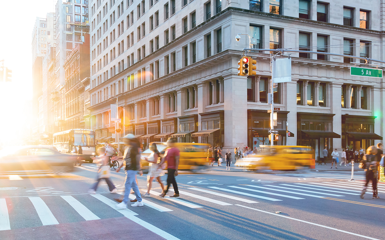 People cross a busy city street with blurred yellow taxis and sunlight streaming in. A large building and traffic lights are visible near the intersection of 5th Ave, capturing the vibrant energy typical of an NYC residential rental neighborhood.