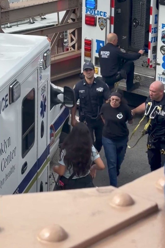 A woman being walked by two police officers, with an ambulance and police truck in the background.
