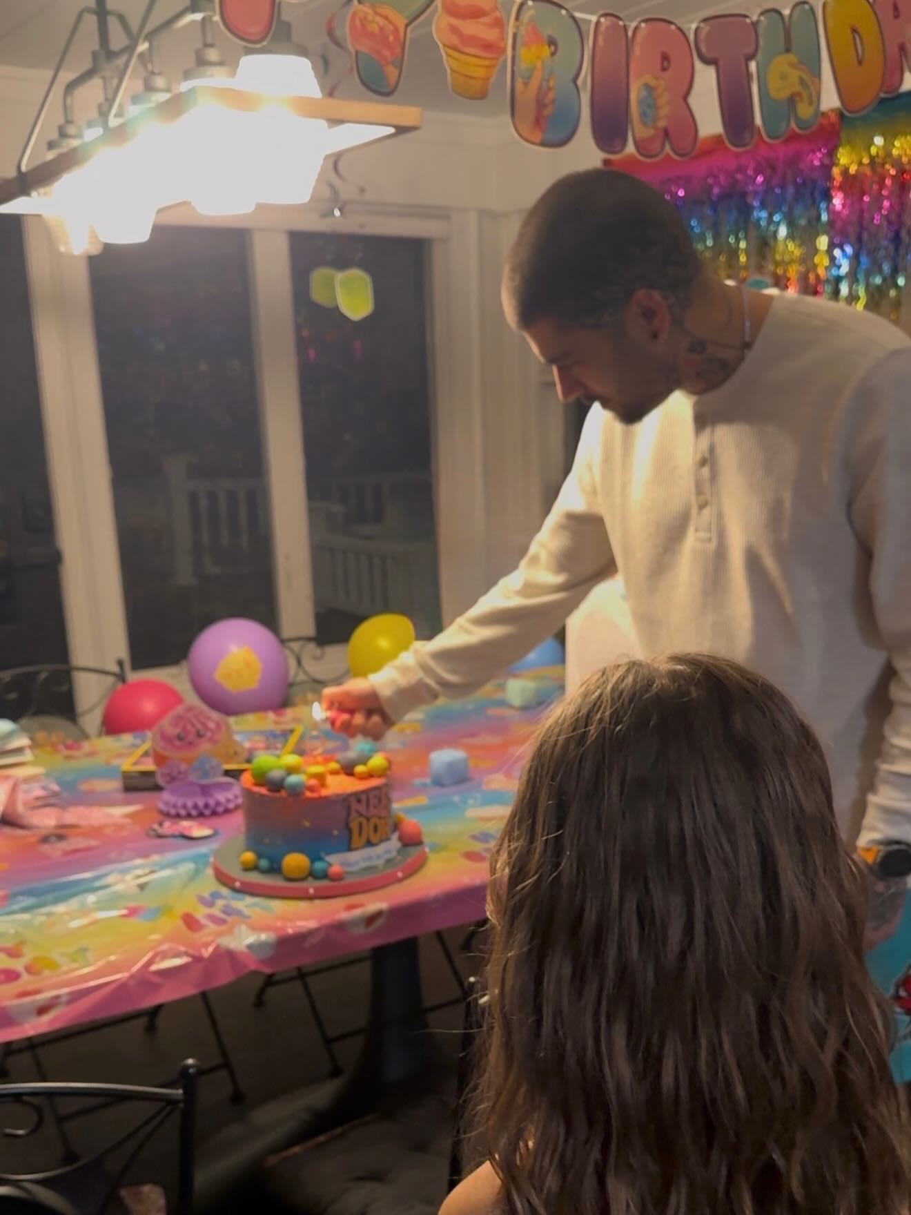 Zayn Malik lighting a birthday candle on a cake, with his daughter in the foreground.