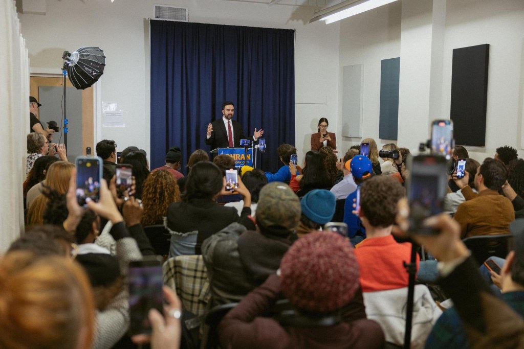 Zohran Mamdani speaks at a news conference to an audience mostly recording on their phones.