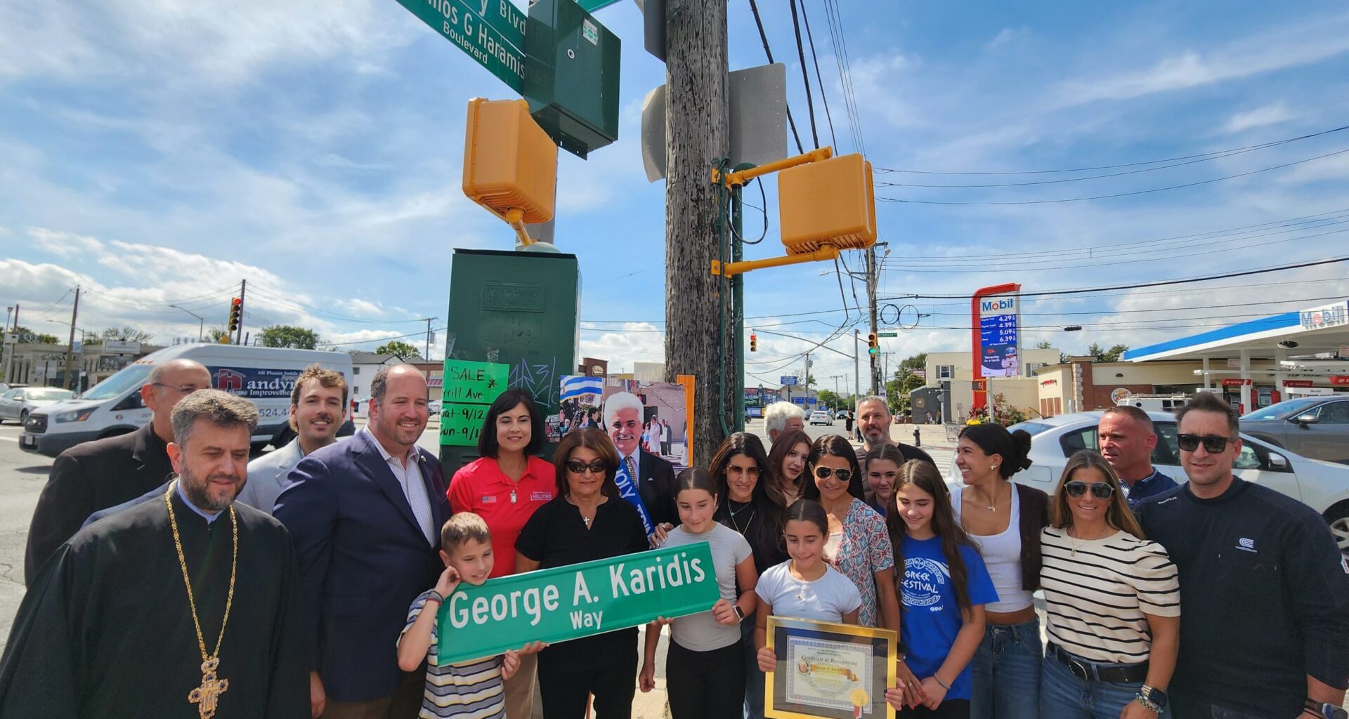 Panagiota Karidis, with her grandchildren, holds a copy of the street sign bearing her husband’s name, which is placed prominently next to the sign for Vassilios Haramis Way, named for a victim of the September 11 attacks. Family members, politicians Nicole Malliotakis and David Carr, and Fr. Vasileios Apostolidis and Fr. Nicholas Anctil can be seen. (Photo: The National Herald/Michael Kakias)