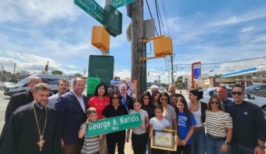 Panagiota Karidis, with her grandchildren, holds a copy of the street sign bearing her husband’s name, which is placed prominently next to the sign for Vassilios Haramis Way, named for a victim of the September 11 attacks. Family members, politicians Nicole Malliotakis and David Carr, and Fr. Vasileios Apostolidis and Fr. Nicholas Anctil can be seen. (Photo: The National Herald/Michael Kakias)